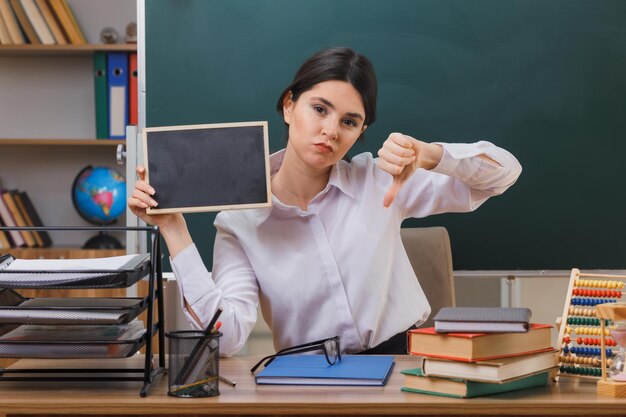 scontento che mostra i pollici verso il basso giovane insegnante femminile che tiene una mini lavagna seduta alla scrivania con gli strumenti della scuola in classe