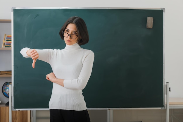 scontento che mostra i pollici verso il basso giovane insegnante femminile con gli occhiali in piedi davanti alla lavagna in classe