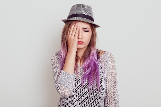 Sconvolto bella donna con i capelli lilla che indossa camicia e cappello essendo triste, che copre metà del viso con il palmo, tiene gli occhi chiusi, isolato sopra il muro bianco.