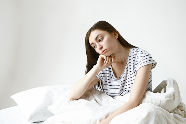 sconvolto bella giovane donna con lunghi capelli castani seduta sul letto, con sguardo pensieroso, riluttante ad andare al lavoro, sentirsi male e stanco della sua noiosa vita monotona