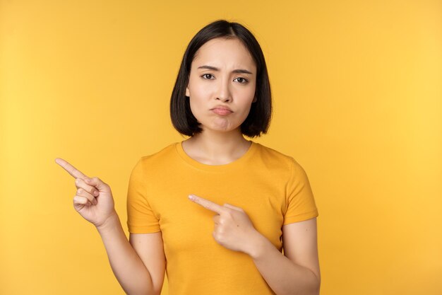 Sconvolto donna asiatica che punta le dita a sinistra mostrando smth deludente lamentandosi al banner in piedi su sfondo giallo