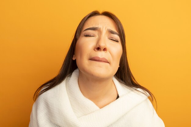Sconvolto giovane donna con sciarpa bianca con gli occhi chiusi con espressione triste in piedi sopra la parete arancione
