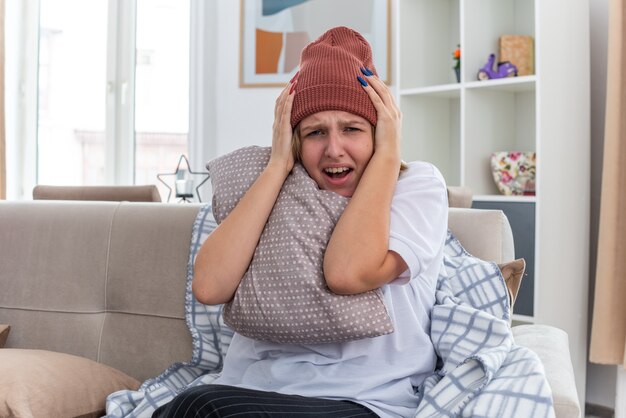 Sconvolto giovane donna malsana in cappello caldo con coperta che sembra malata e malata che soffre di raffreddore e influenza tenendo il cuscino che sembra preoccupata toccando la testa seduta sul divano in un soggiorno luminoso