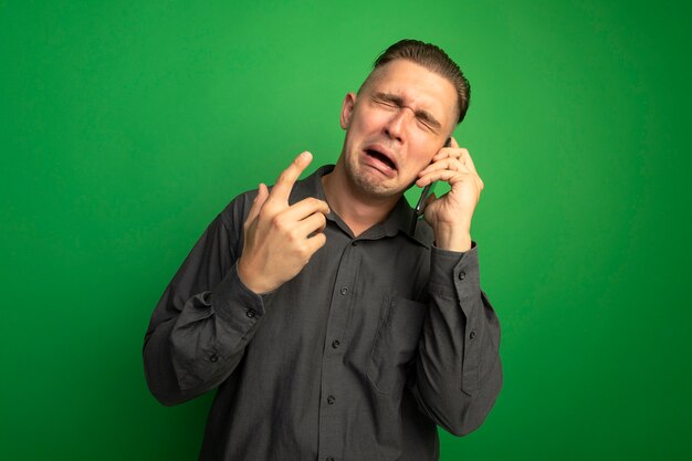 Sconvolto giovane uomo bello in camicia grigia piangere mentre parla al telefono cellulare in piedi sopra la parete verde