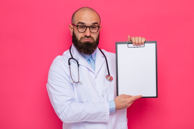 Sconvolto uomo barbuto medico in camice bianco con stetoscopio intorno al collo con gli occhiali che tengono appunti con pagine bianche guardando con espressione triste che increspa le labbra