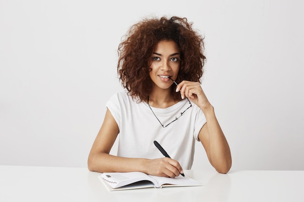 Scrittura di pensiero della ragazza africana in taccuino che sorride sopra la parete bianca. Copia spazio.