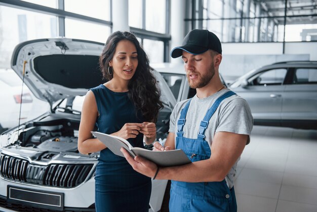 Scrivere usando la penna. Donna nel salone dell'auto con il dipendente in uniforme blu che riprende la sua auto riparata