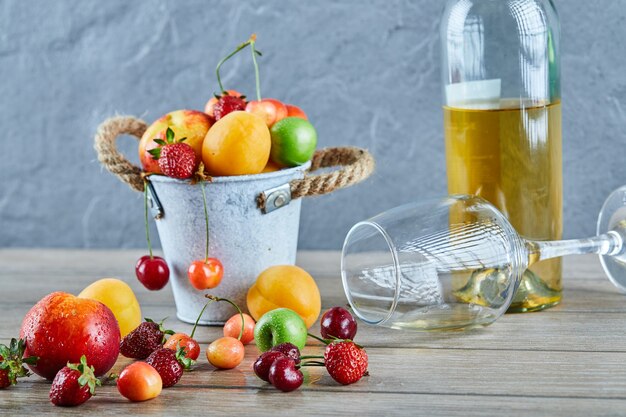 Secchio di frutta fresca estiva, bottiglia di vino bianco e bicchiere vuoto sulla tavola di legno.