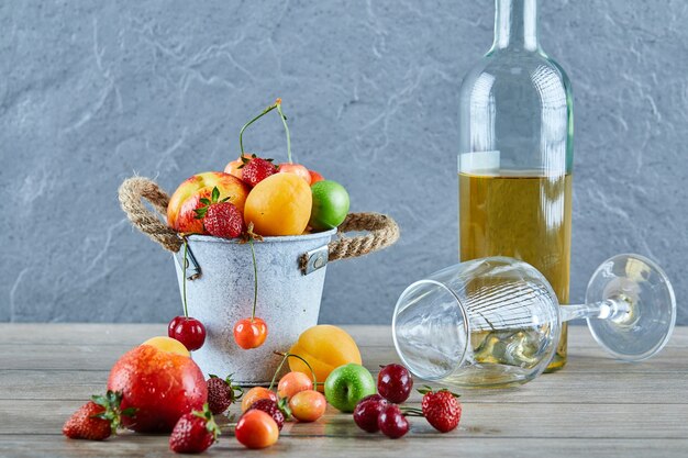 Secchio di frutta fresca estiva, bottiglia di vino bianco e bicchiere vuoto sulla tavola di legno.