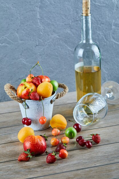 Secchio di frutta fresca estiva, bottiglia di vino bianco e bicchiere vuoto sulla tavola di legno.