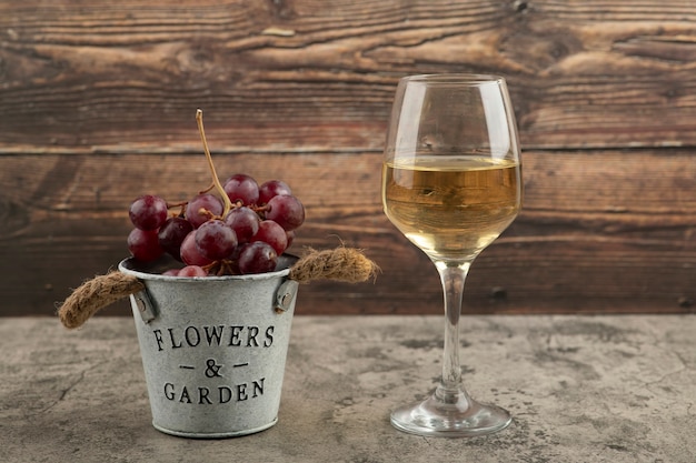 Secchio di metallo di uva fresca rossa e bicchiere di vino bianco sulla superficie di marmo.