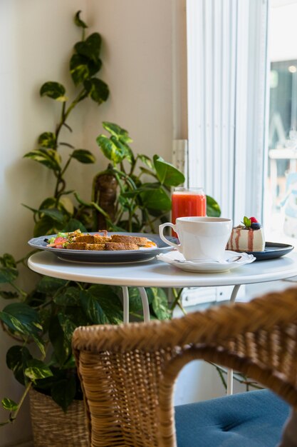 Sedia di vimini vicino al tavolo rotondo bianco con tazza di caffè; prima colazione; frullato e cheesecake