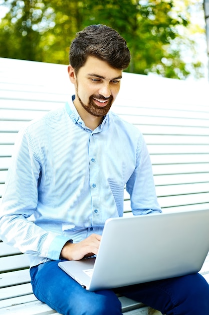Seduta di modello sorridente giovane bello dell'uomo d'affari sul banco di parco facendo uso del computer portatile nel panno casuale dei pantaloni a vita bassa