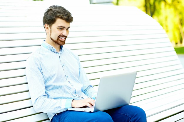 Seduta di modello sorridente giovane bello dell'uomo d'affari sul banco di parco facendo uso del computer portatile nel panno casuale dei pantaloni a vita bassa