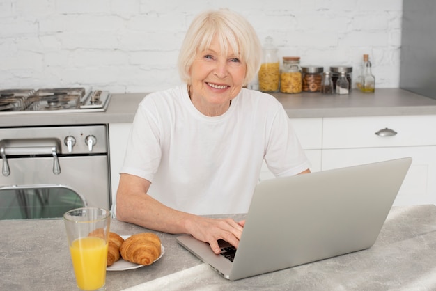Seduta senior felice nella cucina con un computer portatile