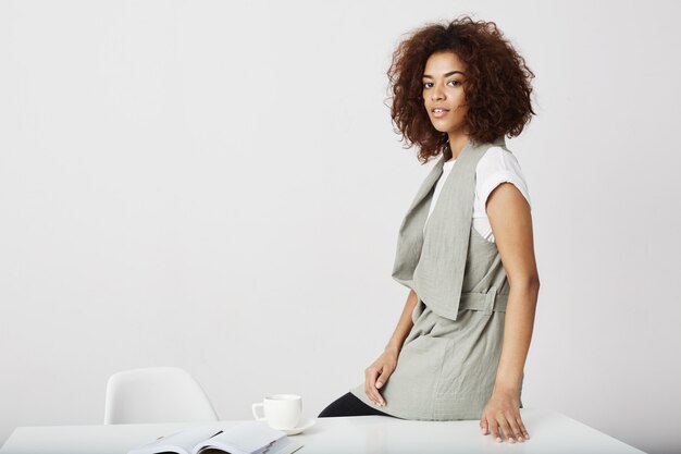 Seduta sorridente della donna di affari africana sulla tavola nel luogo di lavoro sopra la parete bianca.