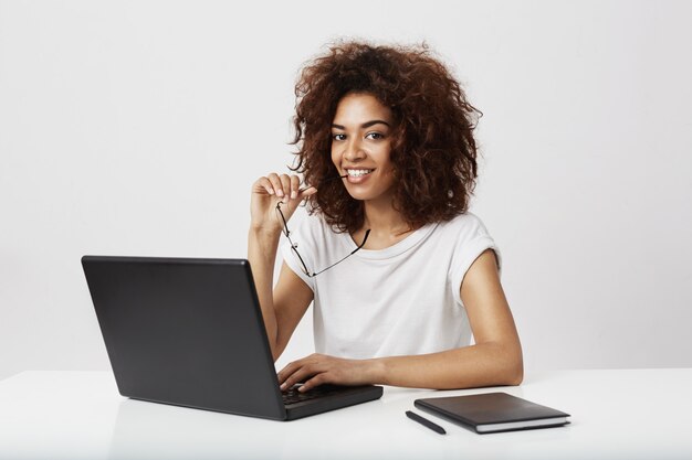 Seduta sorridente della ragazza africana al wokplace con il computer portatile.