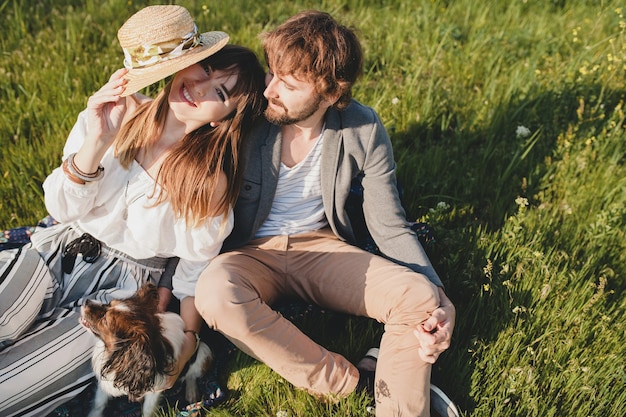 Seduto in erba giovane coppia hipster alla moda innamorata che cammina con il cane in campagna, moda boho stile estivo, romantico