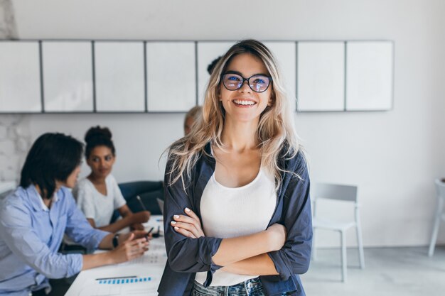 Segretaria femminile soddisfatta in bicchieri alla moda in posa in ufficio dopo l'incontro con i colleghi. Ritratto dell'interno della donna di affari alla moda con i lavoratori asiatici e africani.