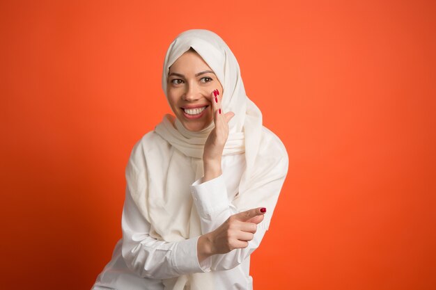 Segreto, concetto di gossip. Felice donna araba in hijab. Ritratto di ragazza sorridente, posa in studio di sfondo rosso. Giovane donna emotiva. Le emozioni umane, il concetto di espressione facciale. Vista frontale.