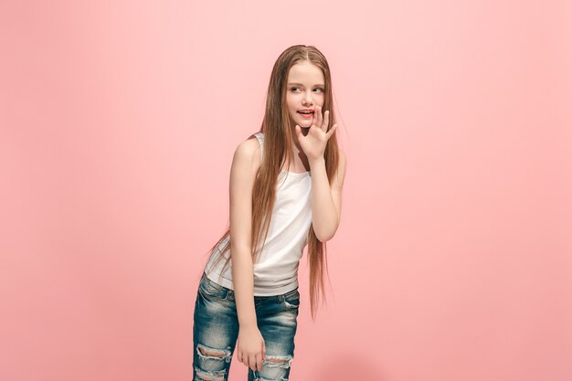 Segreto, concetto di pettegolezzo. Giovane ragazza teenager che bisbiglia un segreto dietro la sua mano isolato su sfondo rosa alla moda per studio.
