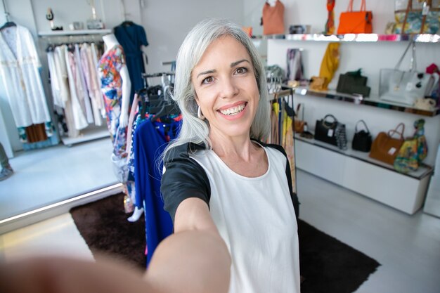 Selfie di donna dai capelli biondi caucasica allegra in piedi vicino a rack con abiti nel negozio di moda, che guarda l'obbiettivo e sorridente Cliente boutique o concetto di assistente di negozio