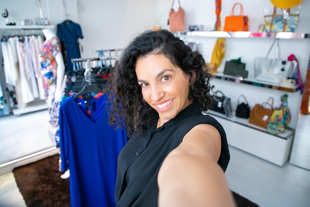 Selfie di donna dai capelli neri latino felice in piedi vicino a cremagliera con abiti nel negozio di moda, che guarda l'obbiettivo e sorridente. Cliente boutique o concetto di assistente di negozio