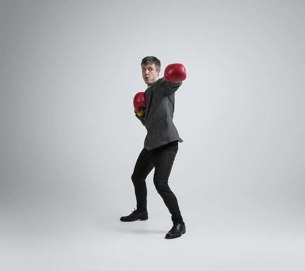 Selvaggio e giovane. Uomo caucasico in abbigliamento da ufficio boxe con due guanti rossi sul muro grigio. Formazione dell'uomo d'affari in movimento, azione. Look insolito per sportivo, attività. Sport, stile di vita sano.