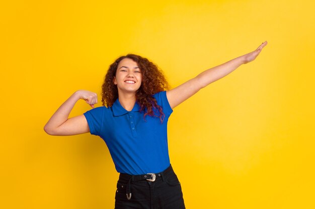 Sembra carino, gesto del vincitore. Ritratto della ragazza dell'adolescente caucasico su sfondo giallo studio. Bello modello riccio femminile in camicia blu. Concetto di emozioni umane, espressione facciale, vendite, annuncio. Copyspace.