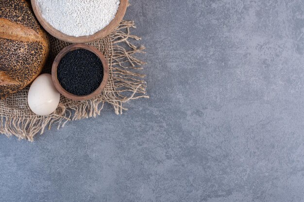 Semi di sesamo neri, farina, uova e una pagnotta di pane rivestita di sesamo su fondo marmo. Foto di alta qualità