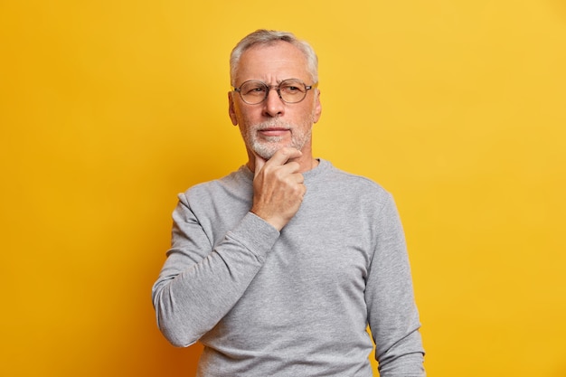 Senior thougthful uomo tiene il mento e guarda pensieroso da parte fa pianificazioni indossa occhiali e casual maglione grigio isolato su vivido muro giallo