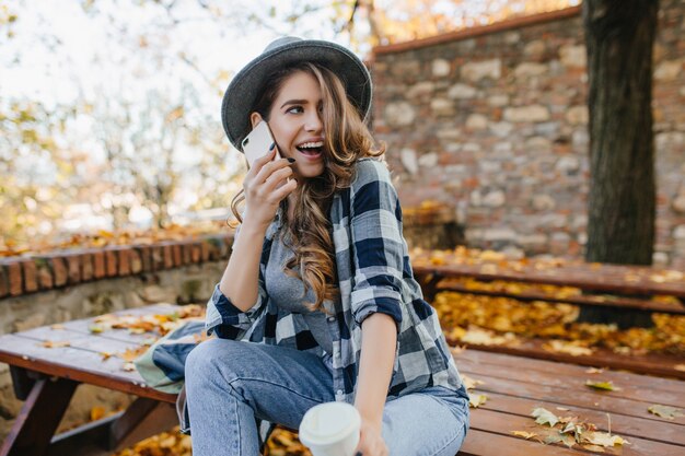 Sensuale donna riccia in cappello che esprime emozioni divertenti durante il servizio fotografico nel cortile di autunno