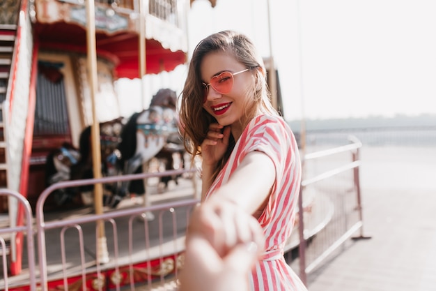 Sensuale giovane modello femminile in posa vicino al carosello. Colpo esterno della ragazza meravigliosa sorridente che gode della giornata nel parco di divertimenti.