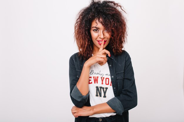 Sensuale ragazza afro-americana con acconciatura riccia alla moda che indossa giacca di jeans e camicia con stampa. Ritratto dell'interno dell'affascinante modello femminile nero in posa giocosamente.