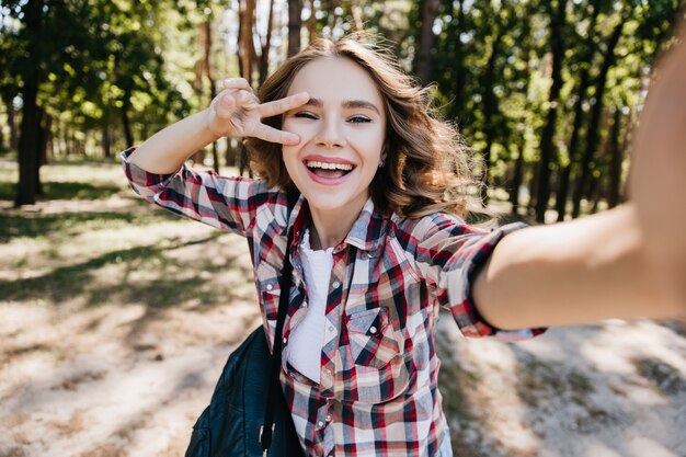 Sensuale ragazza bianca con zaino in posa con il segno di pace nella foresta. Colpo esterno della splendida donna riccia che fa selfie nel parco e ridendo.