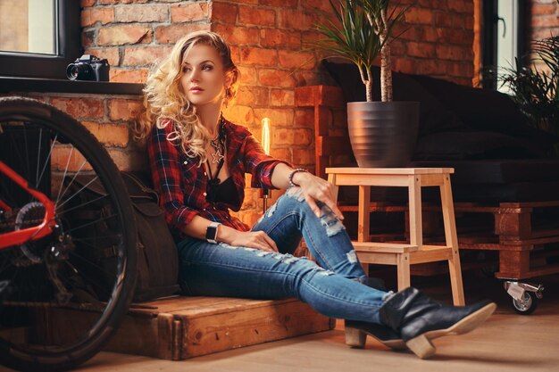 Sensuale ragazza bionda hipster con lunghi capelli ricci vestita con una camicia in pile e jeans seduta su una scatola di legno e appoggiata su un muro di mattoni, guardando lontano, in uno studio con interni soppalcati.