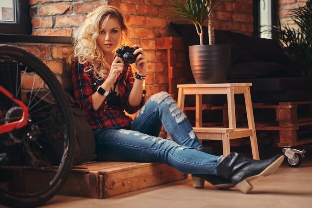 Sensuale ragazza bionda hipster con lunghi capelli ricci vestita con una camicia in pile e jeans tiene una macchina fotografica seduta su una scatola di legno in uno studio con interni soppalcati, guardando una macchina fotografica.