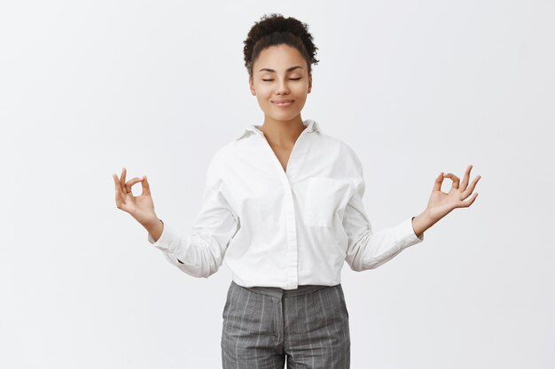 Senza stress, solo pace dentro. Affascinante donna rilassata e spensierata in abito prepotente, alzando le mani in gesto zen, sorridendo con gli occhi chiusi mentre medita o pratica yoga, sentendosi sollevata