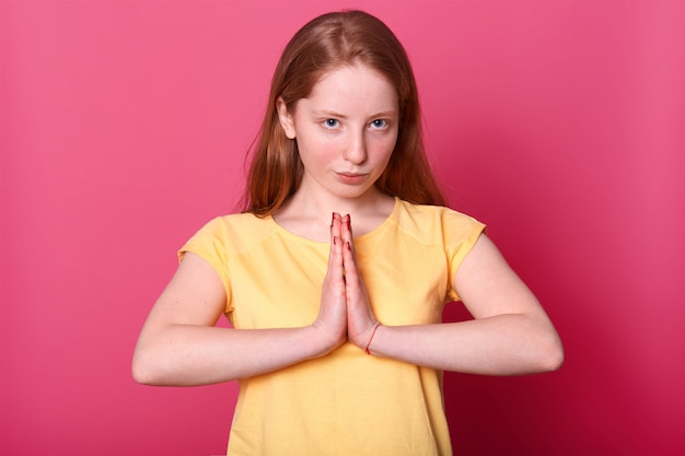 seria femmina caucasica in posa con espressione fiduciosa, tiene le mani in gesto di preghiera, indossa maglietta gialla, isolata sul rosa.