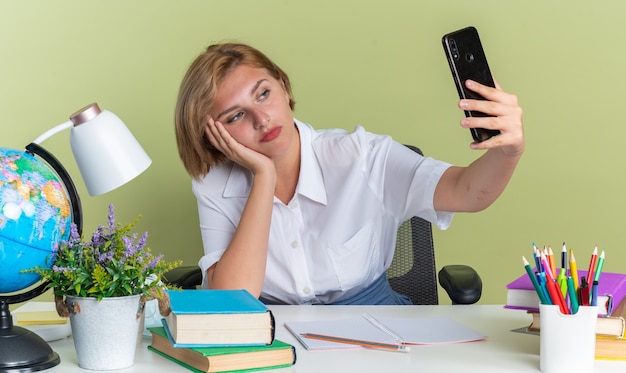 Seria giovane studentessa bionda seduta alla scrivania con gli strumenti della scuola che tiene la mano sul viso prendendo selfie isolata sul muro verde oliva