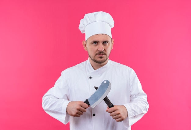 Seriamente cercando giovane bello cuoco in uniforme da chef tenendo la mannaia e coltello isolato su spazio rosa