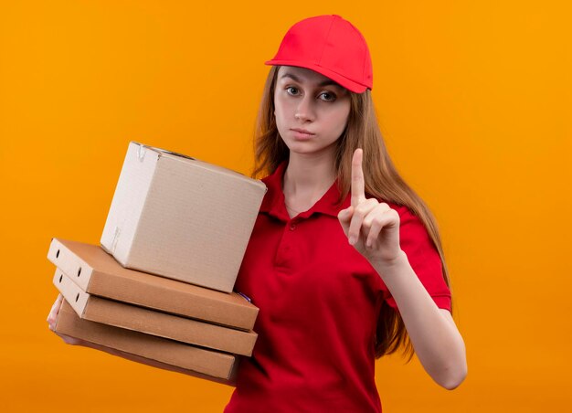 Seriamente cercando giovane ragazza di consegna in rosso uniforme che tiene scatola e pacchetti con il dito alzato su uno spazio arancione isolato