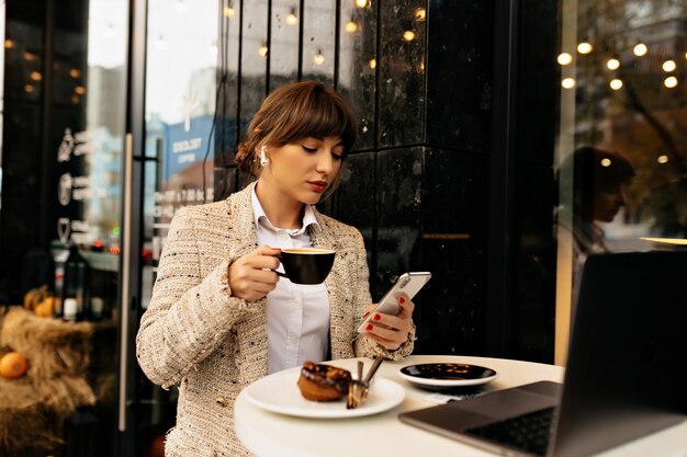 Seriamente giovane elegante donna d'affari in giacca utilizza lo smartphone e lavora al computer portatile, ascoltando musica e avendo pausa caffè