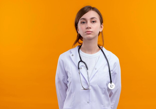 Seriamente guardando giovane medico femminile che indossa abito medico e stetoscopio guardando isolato spazio arancione con copia spazio