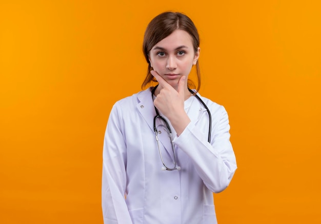 Seriamente guardando giovane medico femminile che indossa abito medico e stetoscopio mettendo la mano sul mento sullo spazio arancione isolato con lo spazio della copia