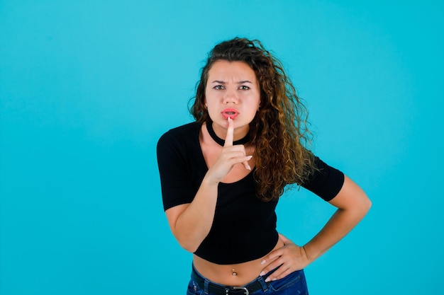 Seriamente la ragazza sta mostrando il gesto del silenzio tenendo l'indice sulle labbra su sfondo blu