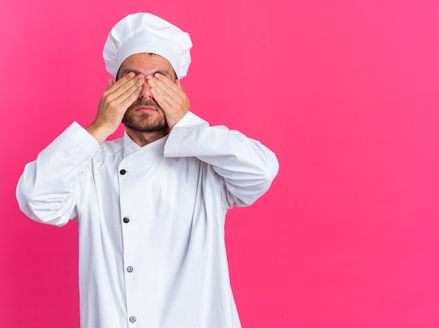 Serio giovane maschio caucasico cuoco in uniforme da chef e berretto che copre gli occhi con le mani isolate sulla parete rosa con spazio copia