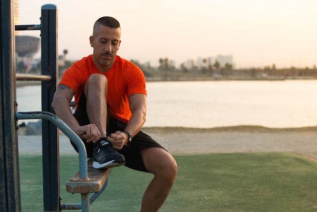 Serio giovane uomo legare le scarpe da ginnastica sul parco giochi