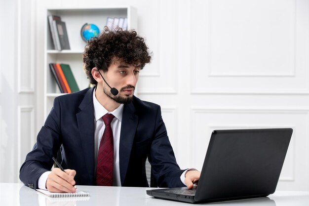 Servizio clienti bel ragazzo in tuta da ufficio con laptop e auricolare scrivendo note