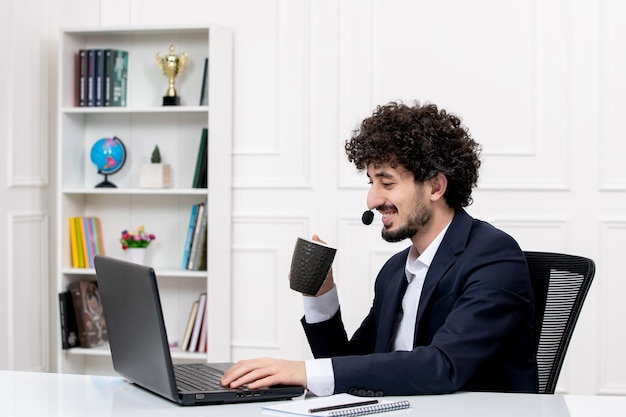 Servizio clienti bell'uomo riccio in tuta da ufficio con computer e auricolare che beve caffè